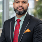 A professional portrait of Akram Hossan wearing a black suit jacket, red tie, and orange pocket square.